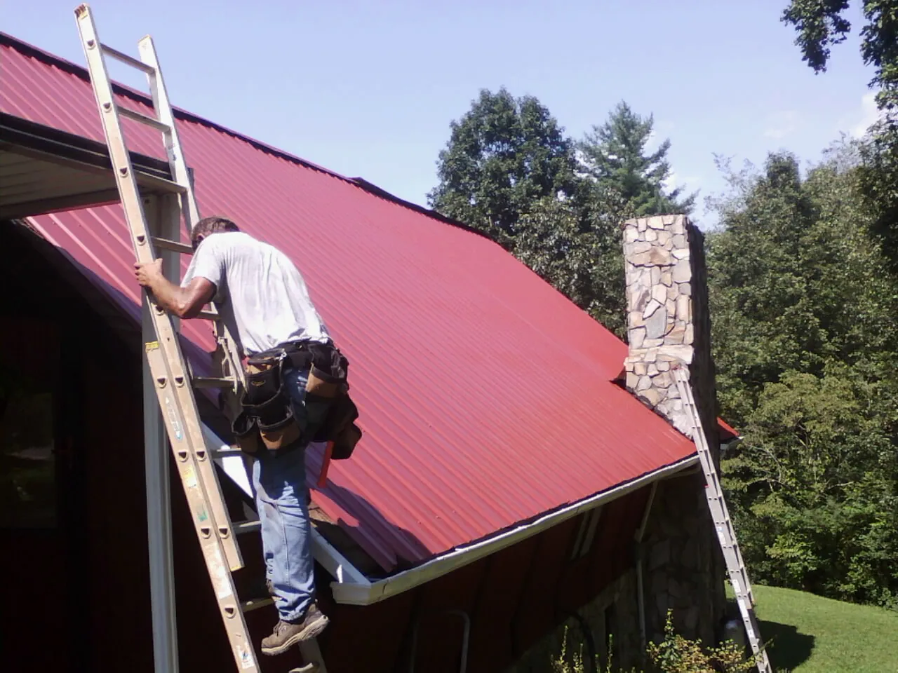 Roofer on a ladder beside a red metal roof and stone chimney — Soffit & Fascia Repair by Roofing Brothers in Alex