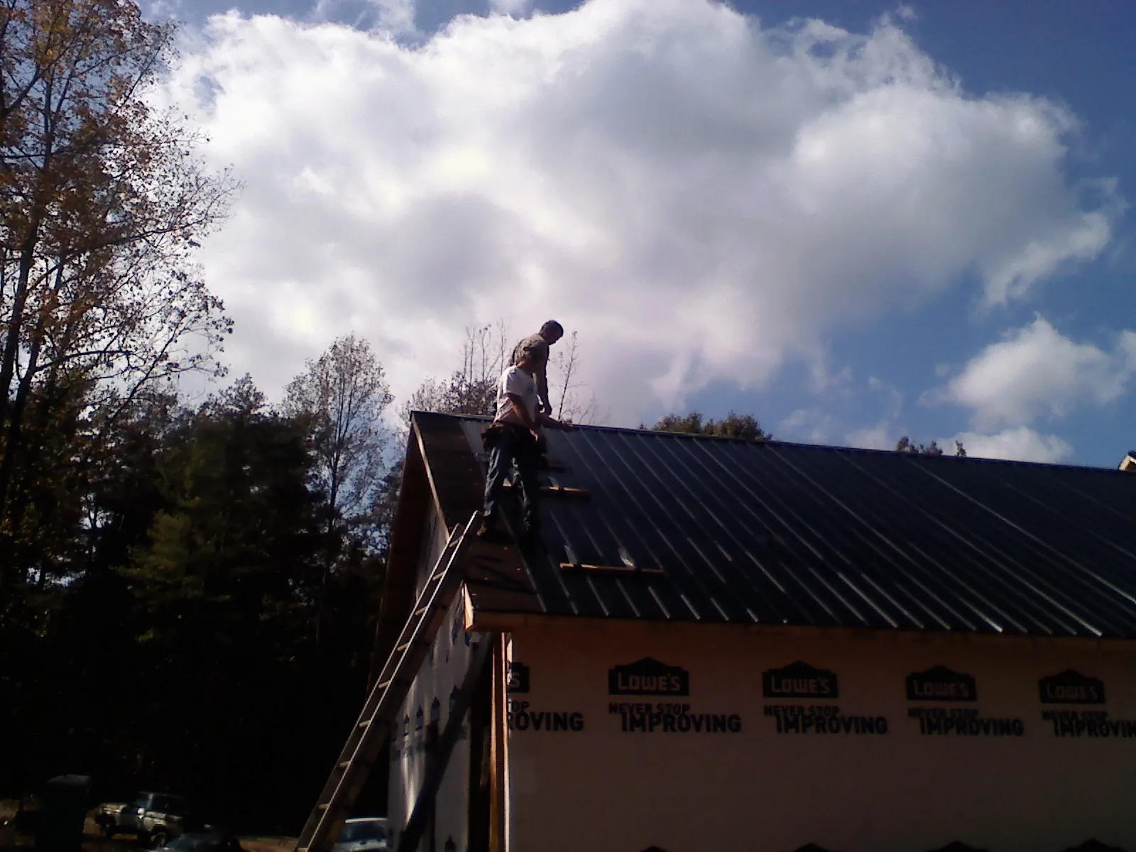 Roofer fastening dark metal panels high against a cloudy sky, from a Roof Inspection job in Alex